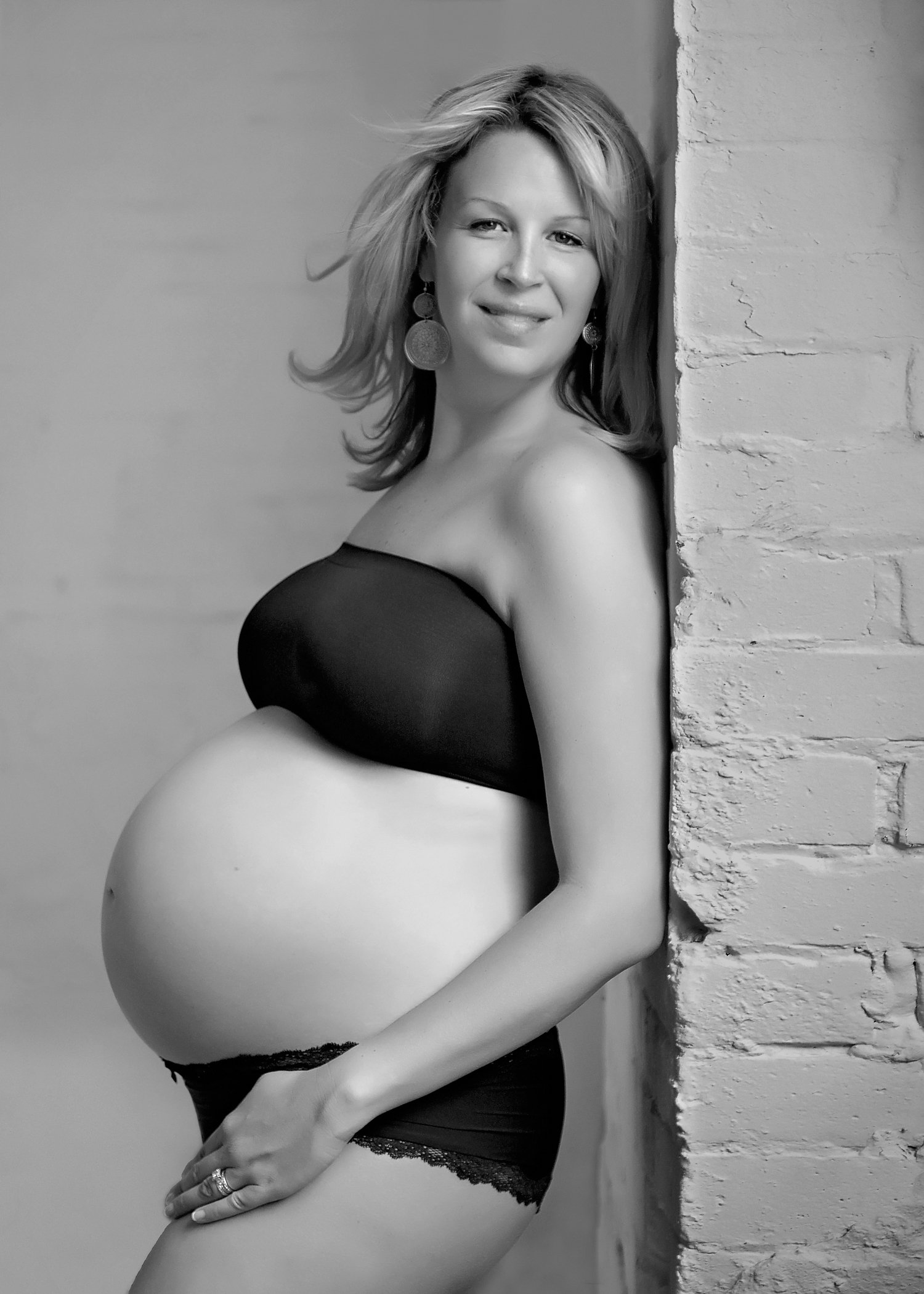 A relaxed pregnant woman leaning against a brick wall, wearing black lingerie and smiling.