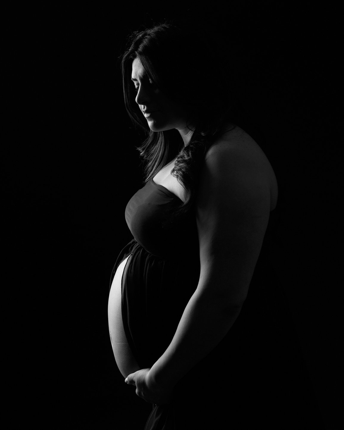 Pregnant woman in a black dress, captured in a dramatic silhouette against a dark background. Toronto maternity portraits