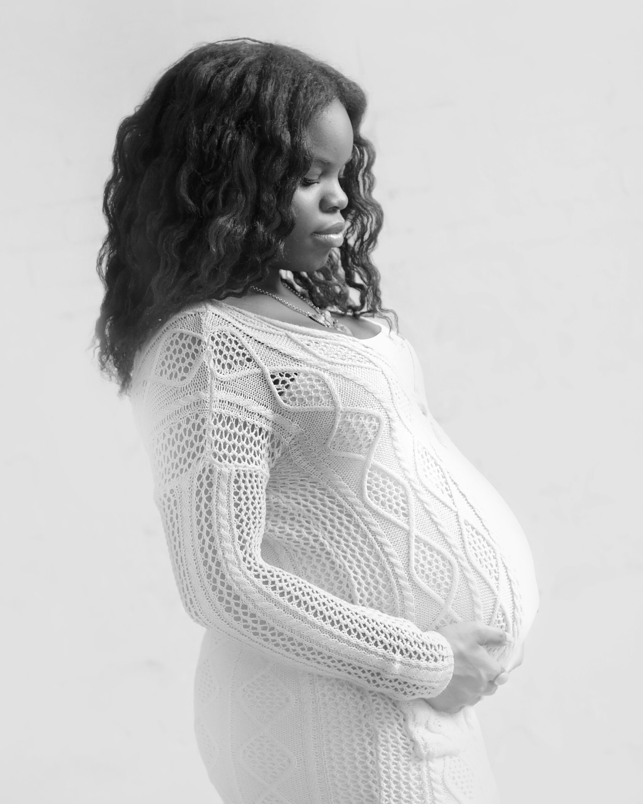 A serene portrait of a pregnant woman in a white dress, standing sideways and looking down at her belly