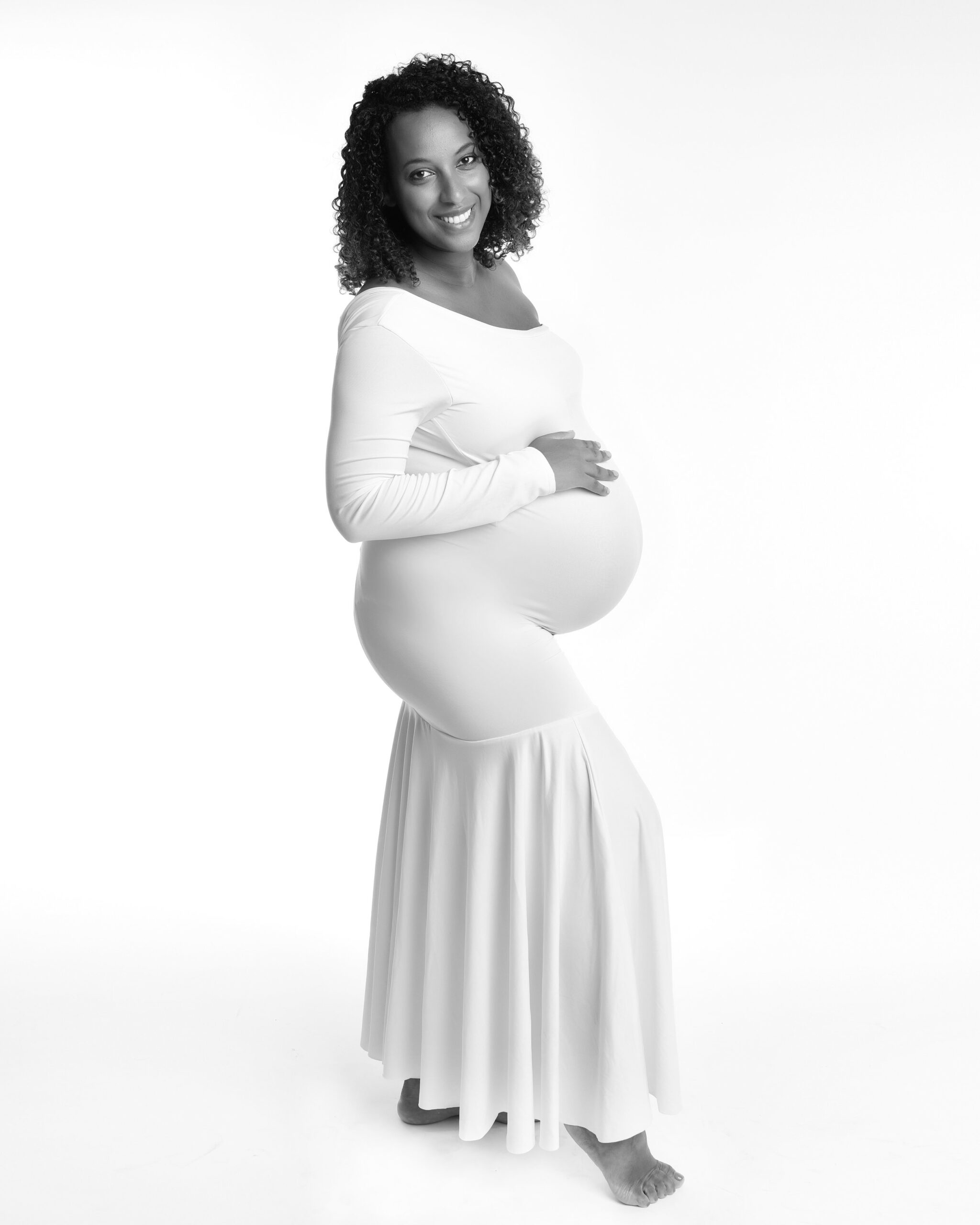 Natural maternity photography TorontoSmiling pregnant woman in a flowing white dress, standing and cradling her belly in a bright studio. A toronto maternity photography photo shoot