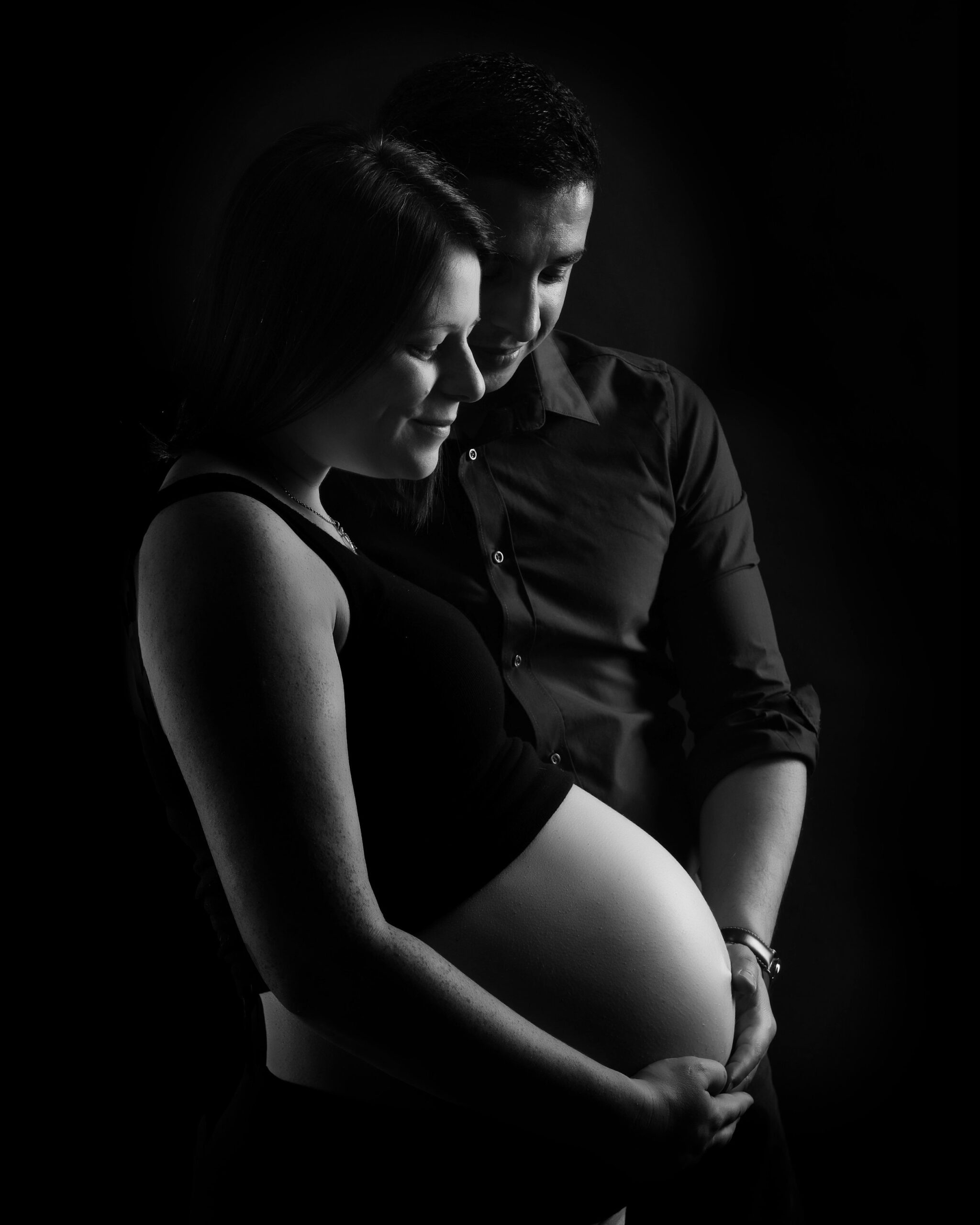 Expectant couple embracing, with the man's hands gently resting on the woman's pregnant belly, captured in a loving black and white portrait. Toronto maternity photo sessions