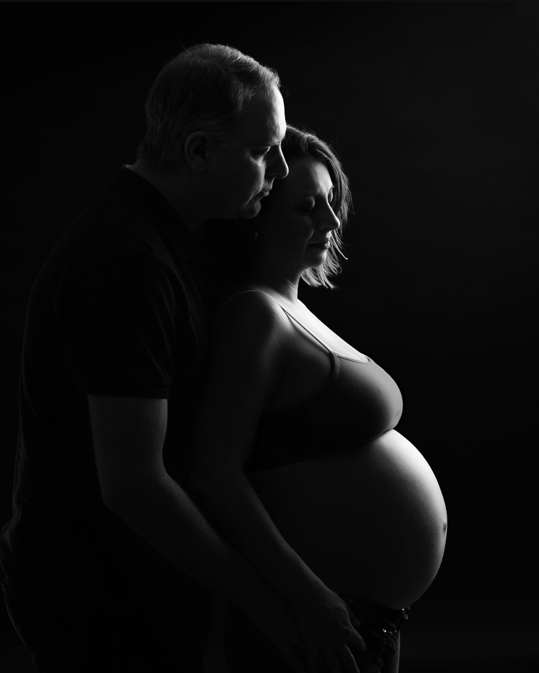 Expectant couple embracing, with the man's face close to the woman's, creating an intimate and loving black and white portrait. Toronto baby bump photography