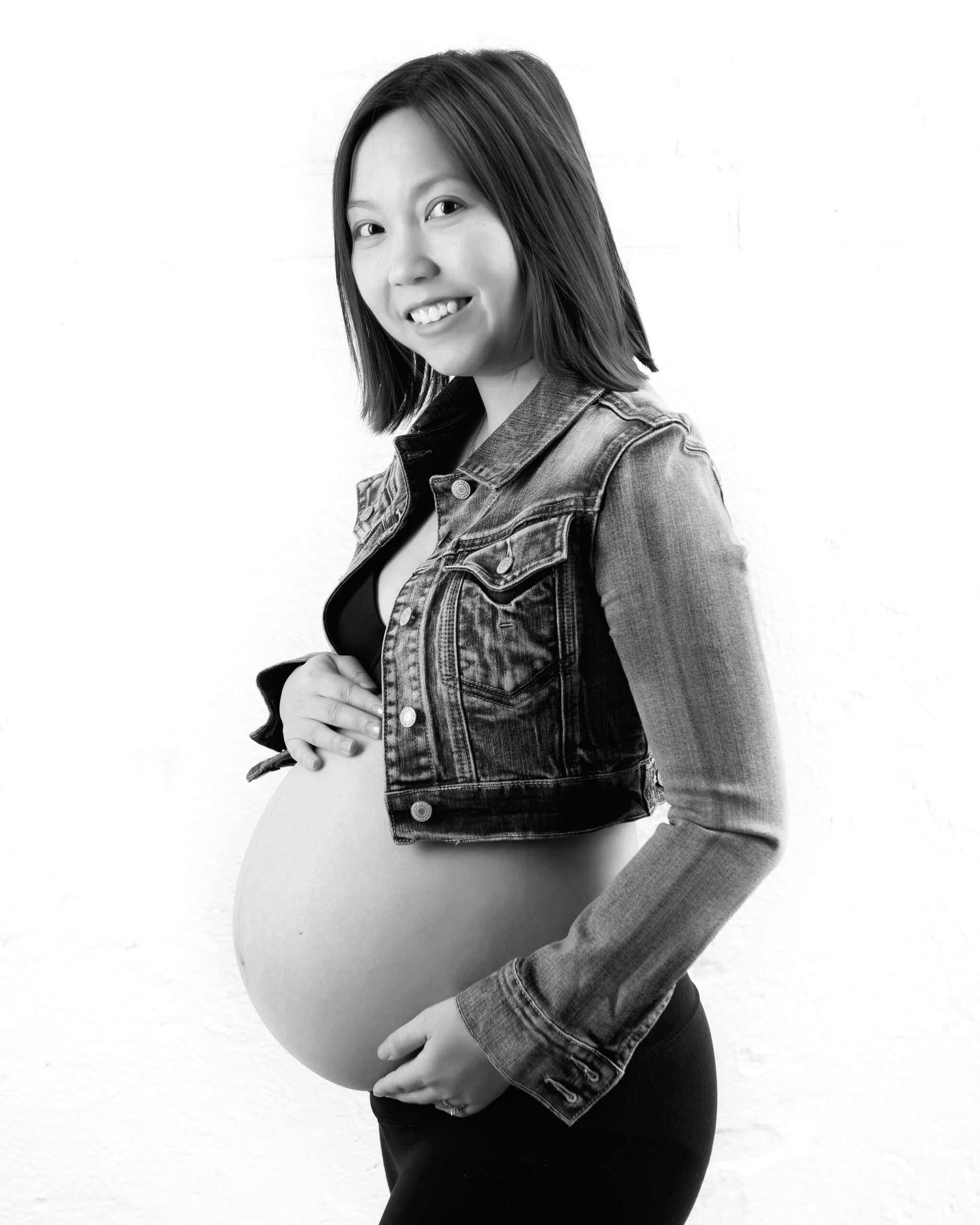 Pregnant woman wearing a denim jacket and black leggings, smiling and holding her belly in a modern studio setting. Toronto expectant mother photography