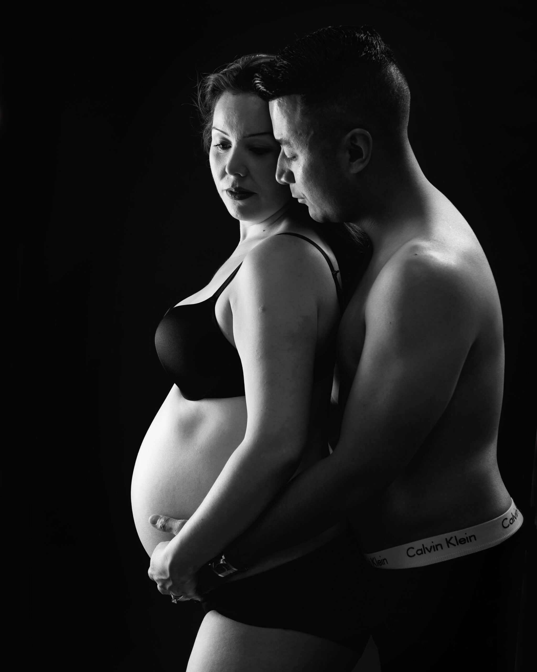 A couple stands closely, with the man embracing the woman from behind, both hands on her pregnant belly. They are both wearing black, set against a dark background, creating an intimate and loving portrait.