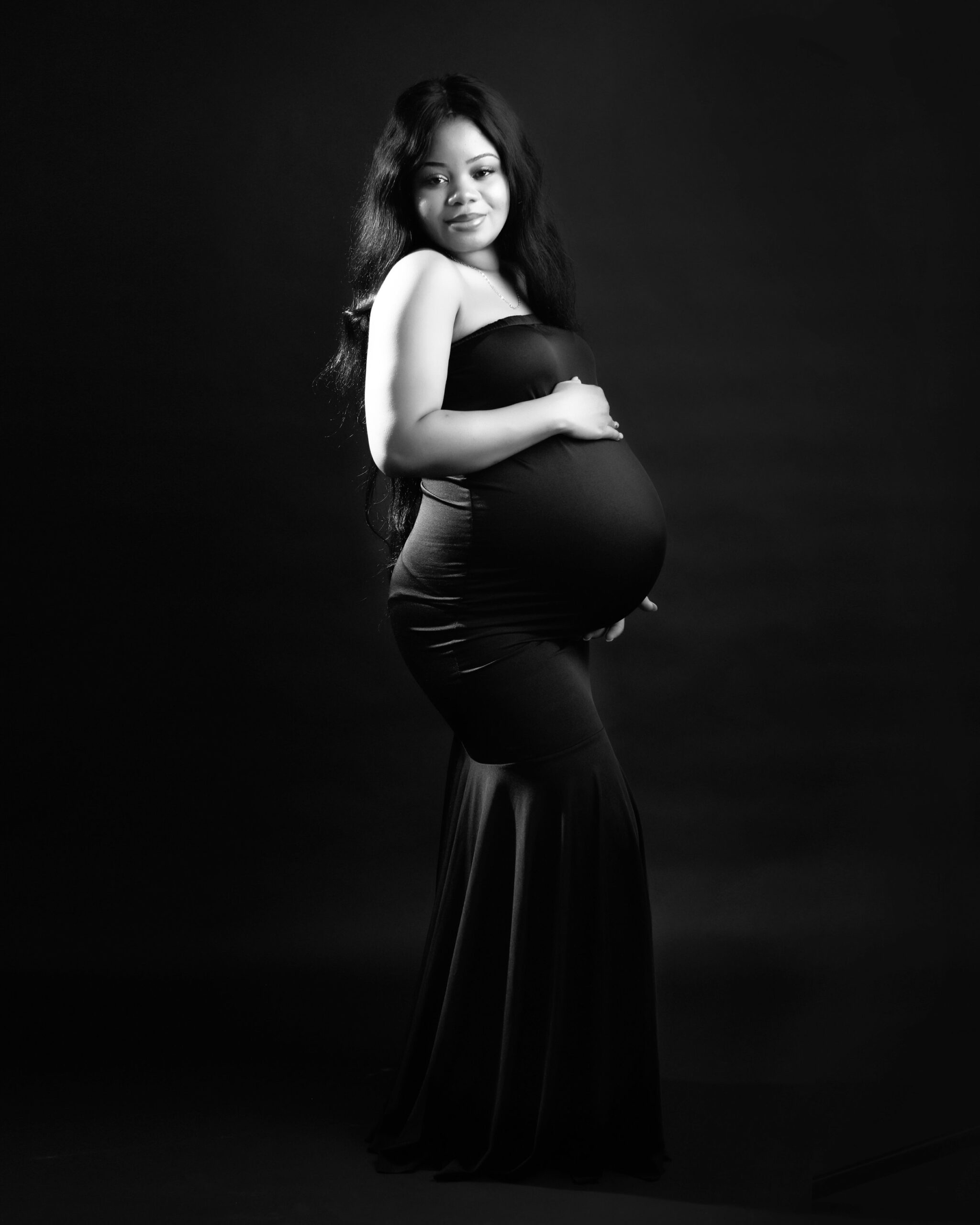 A pregnant woman in a black gown stands with a hand on her belly, her long hair flowing over her shoulders. The image is set against a dark background, giving it a timeless feel.