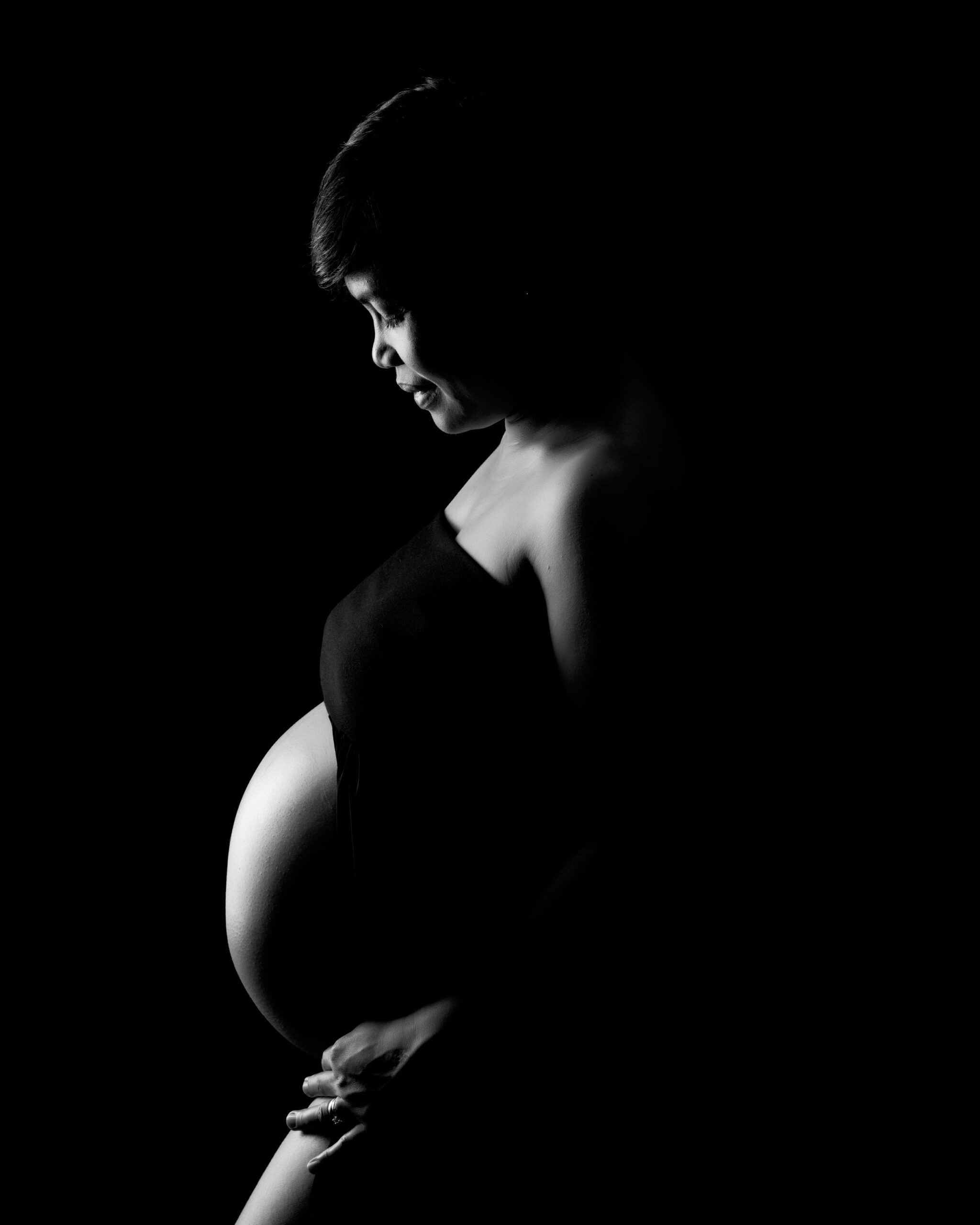 Profile silhouette of a pregnant woman in a black dress, highlighting her belly against a dark background. Toronto motherhood photography