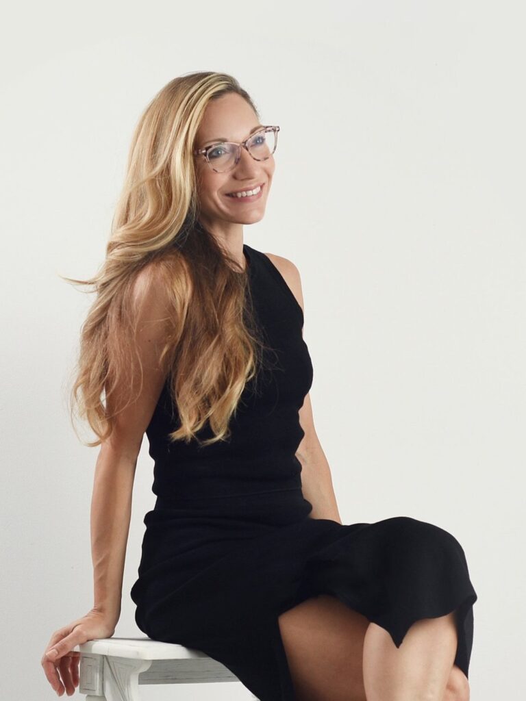 Carla Morgado with long blonde hair wearing glasses and a black dress, sitting gracefully on a white chair. photographers toronto