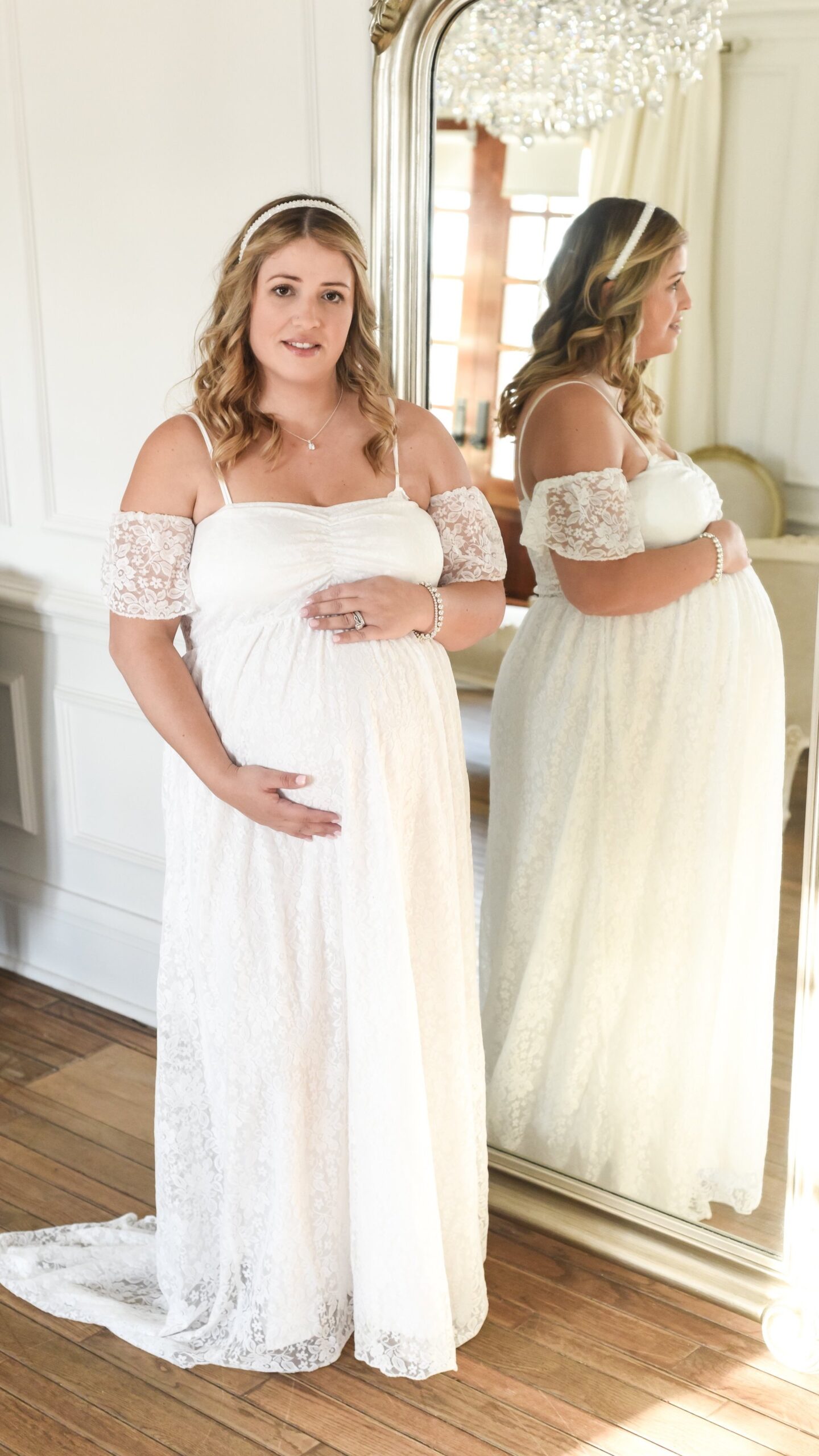 An elegant pregnant woman in a white lace dress, standing in front of a mirror and gently holding her belly. A toronto maternity photography photo shoot