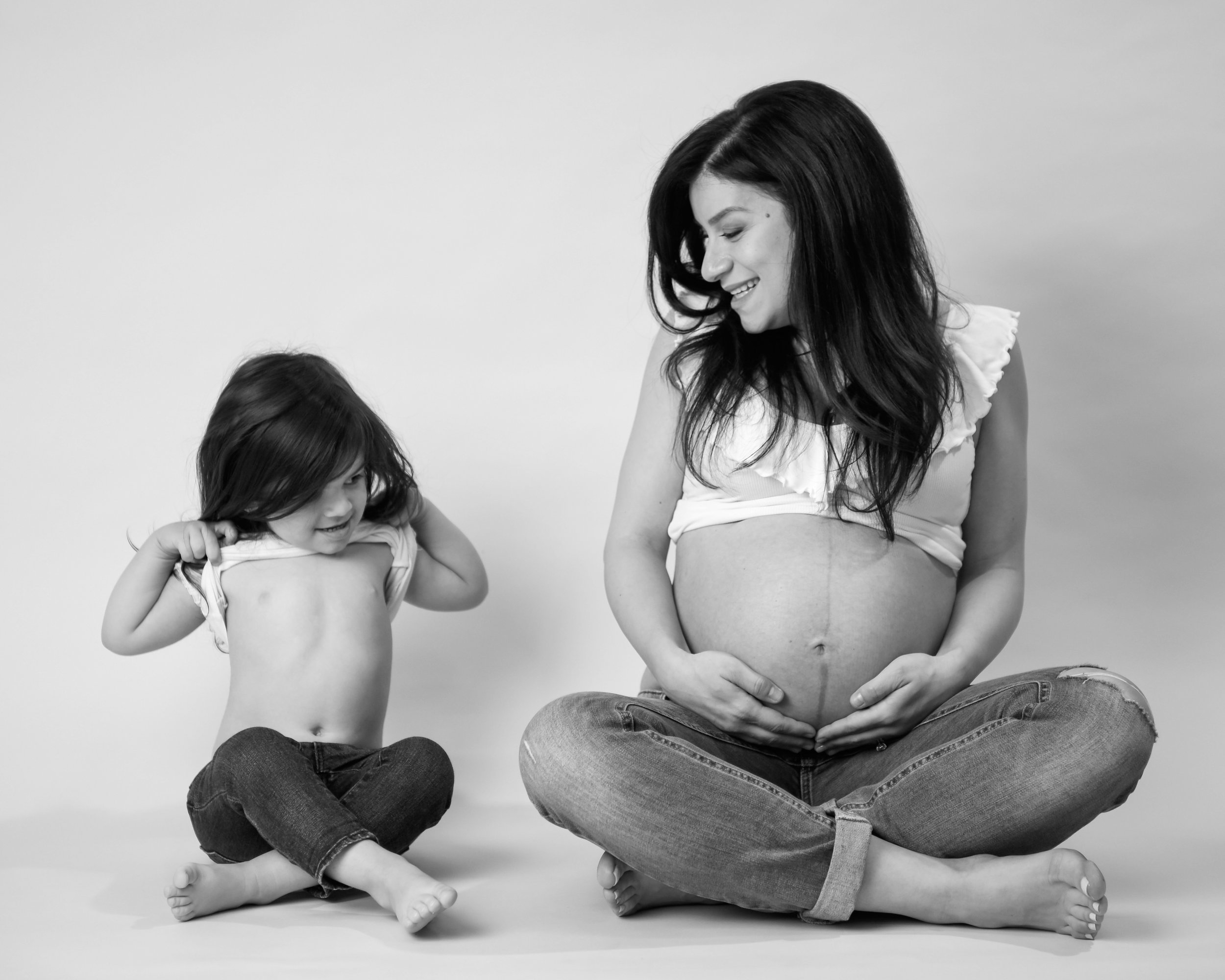 A pregnant woman and her young daughter, both wearing jeans and white tops, sit on the floor laughing and lifting their shirts to reveal their bellies. The image captures a playful and loving moment. A toronto maternity photography photo shoot