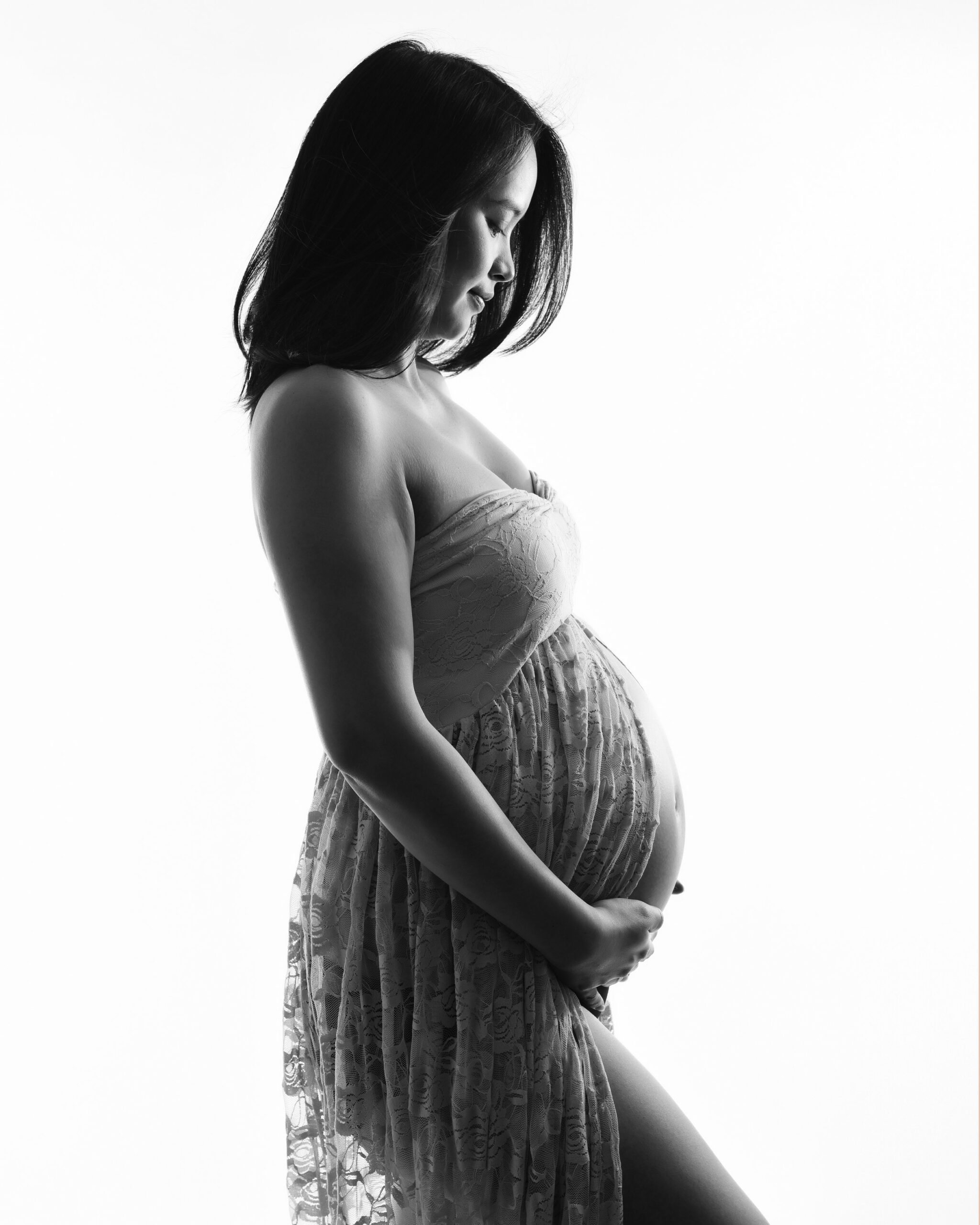 A pregnant woman in a sheer lace dress stands in profile, cradling her belly. The high-key lighting creates an ethereal silhouette effect, emphasizing her peaceful expression. A toronto maternity photography photo shoot

