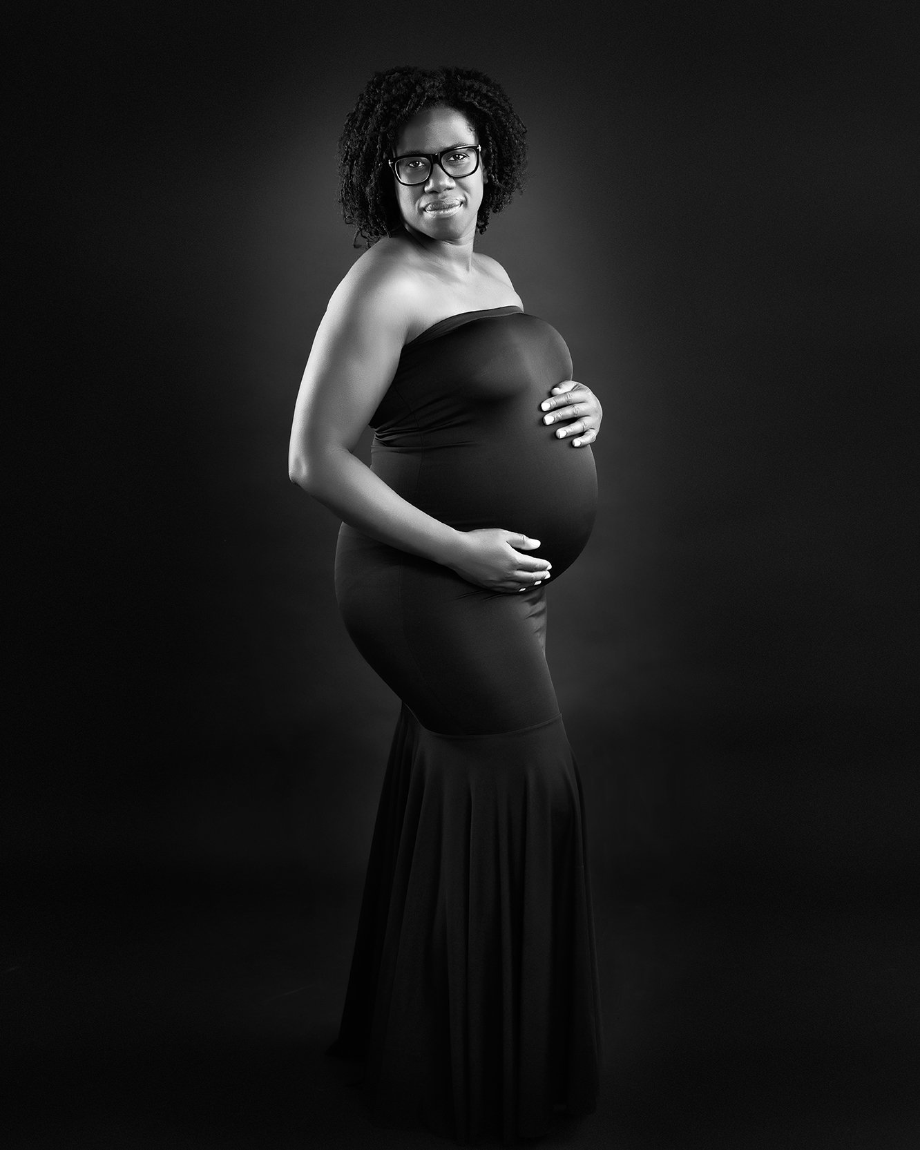 A stylish portrait of a pregnant woman in a black dress, standing and smiling at the camera.