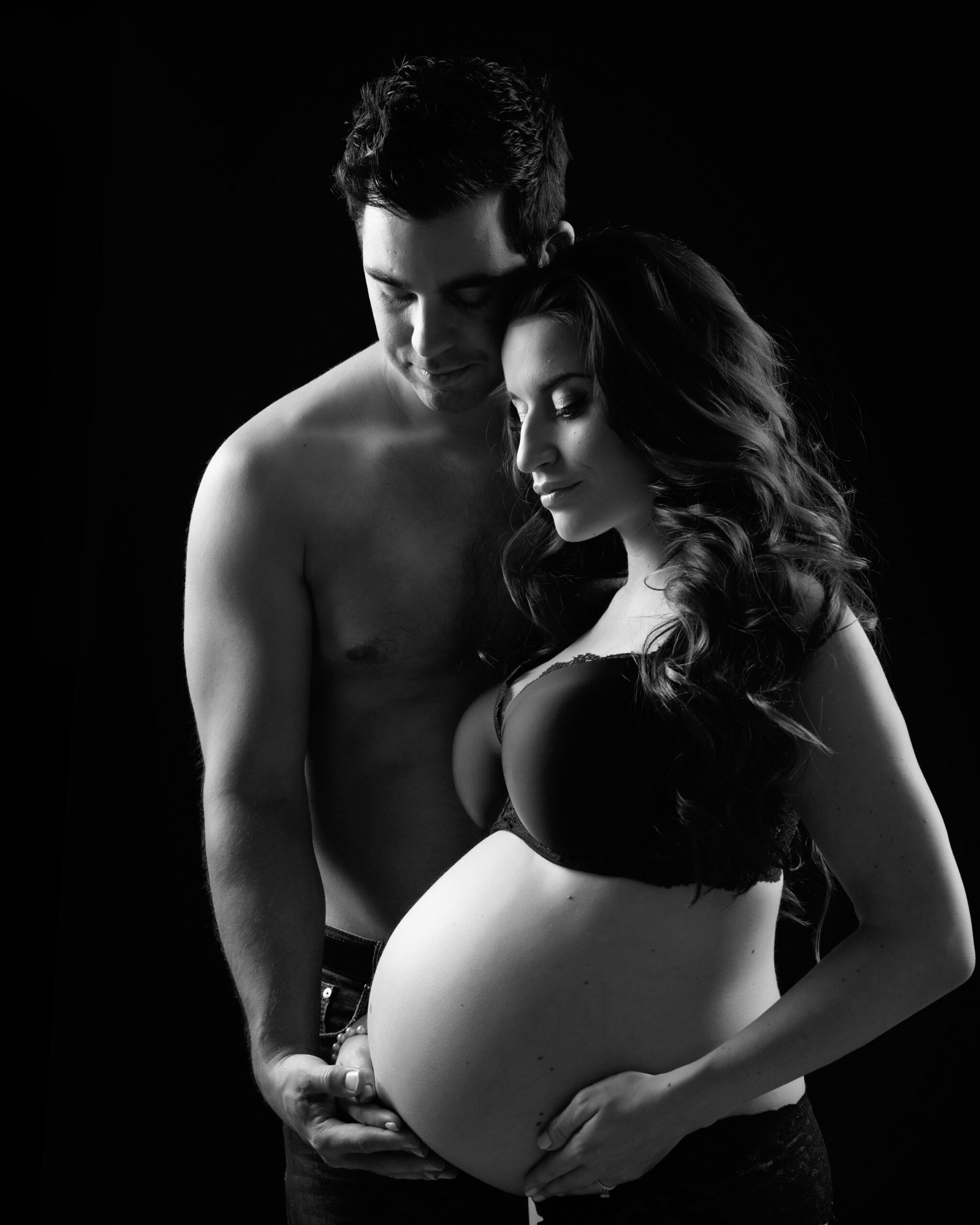 A couple embraces tenderly with the man holding the woman's pregnant belly. They are both shirtless and posed against a dark background, creating a dramatic and intimate portrait.