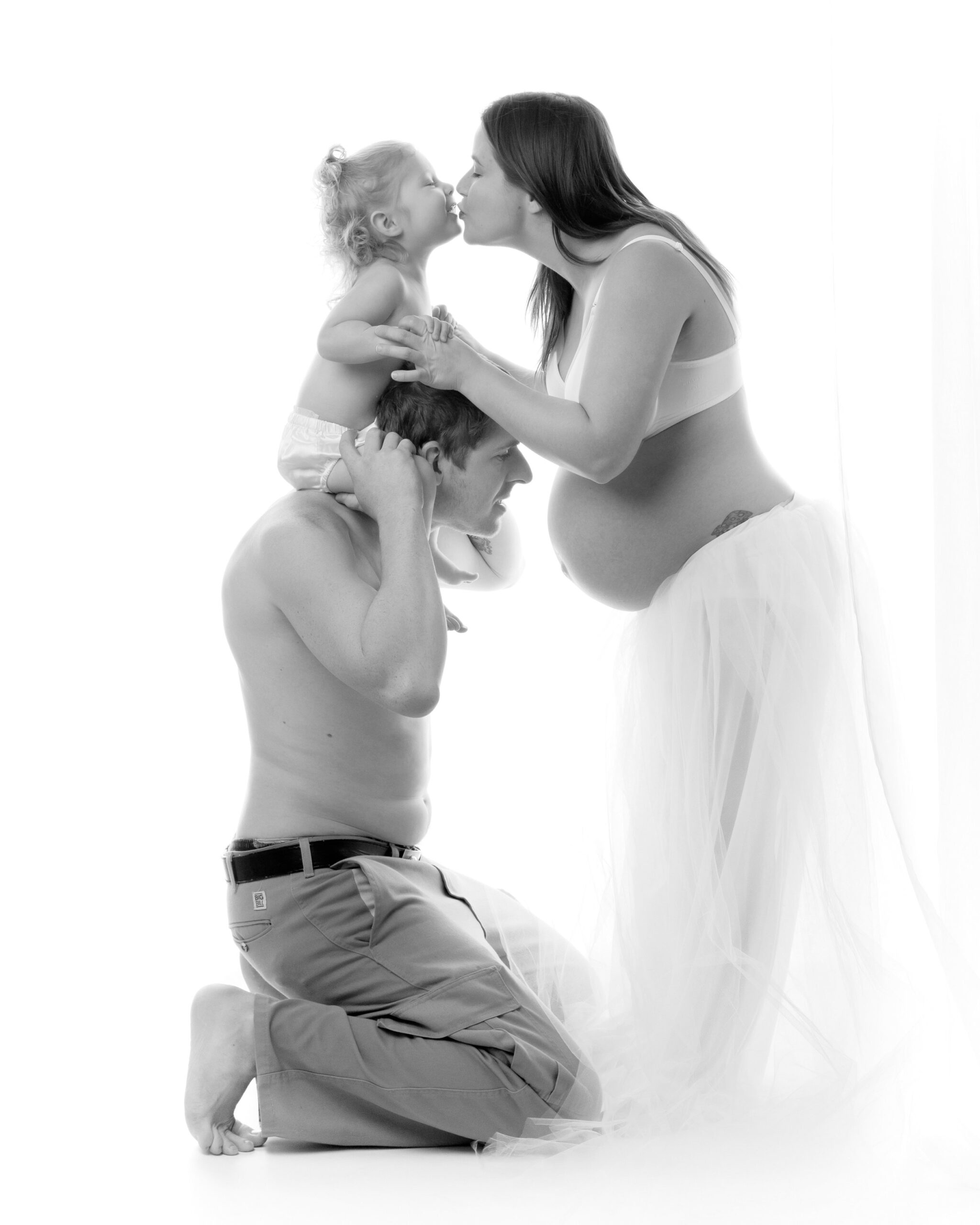 A pregnant woman in a white outfit kisses her young daughter who is sitting on her father's shoulders. The family is posed against a bright white background. A toronto maternity photography photo shoot. 