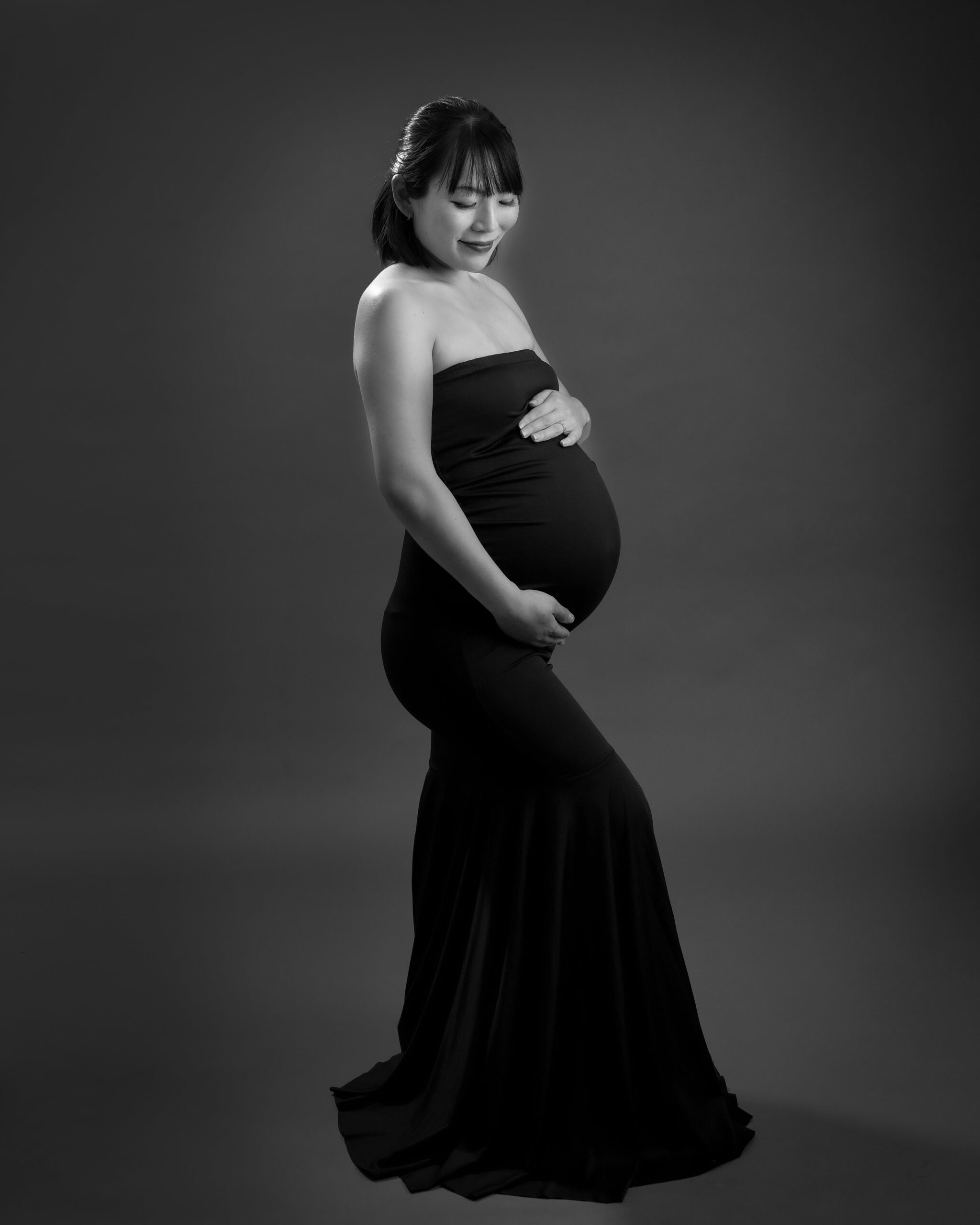 A pregnant woman in a form-fitting black gown poses gracefully, holding her belly with both hands. The image has a dark, elegant background, highlighting her serene expression.