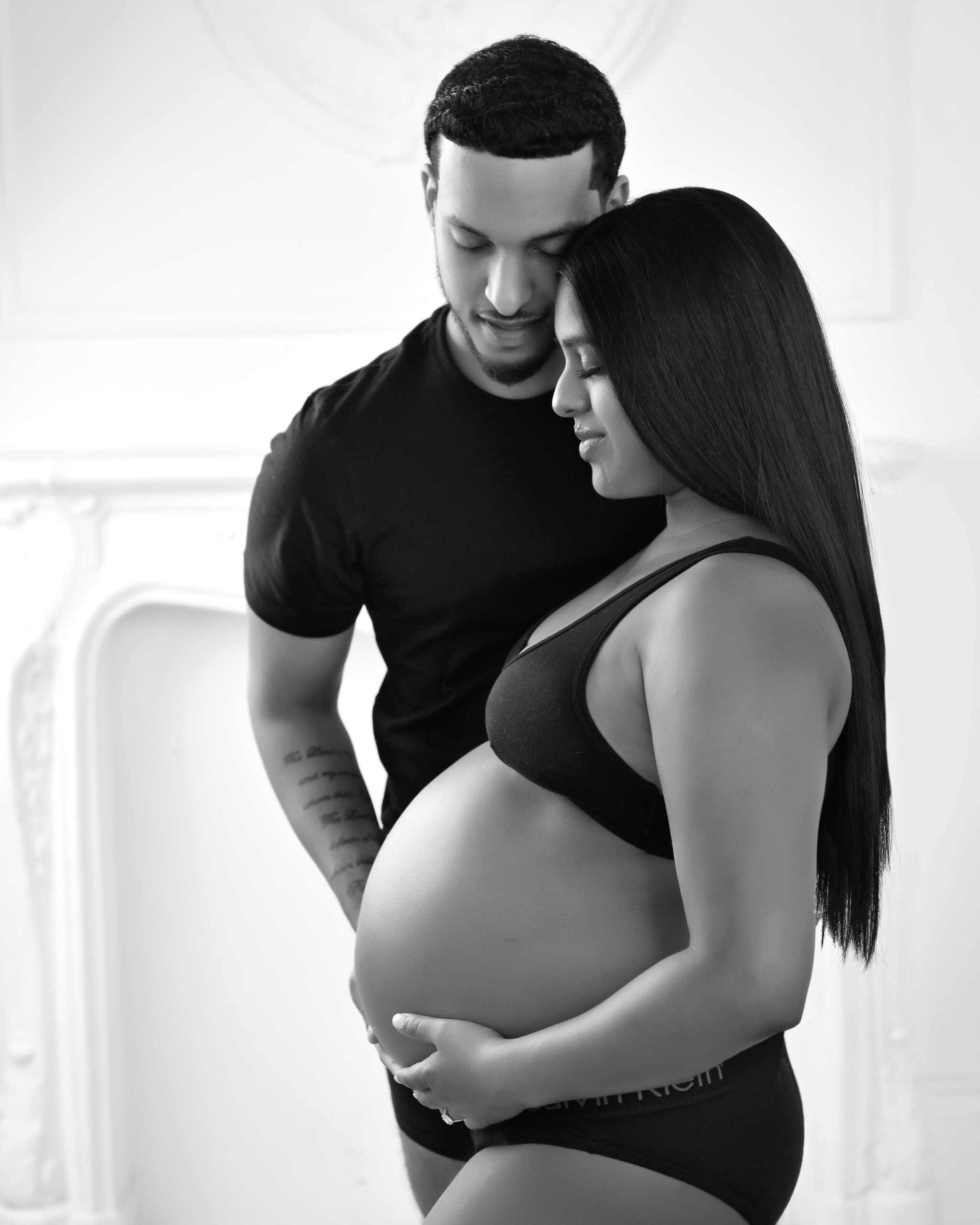 A couple stands close together, the woman cradling her pregnant belly while her partner gently touches it. Both are dressed in black, and the image is taken against a bright background.