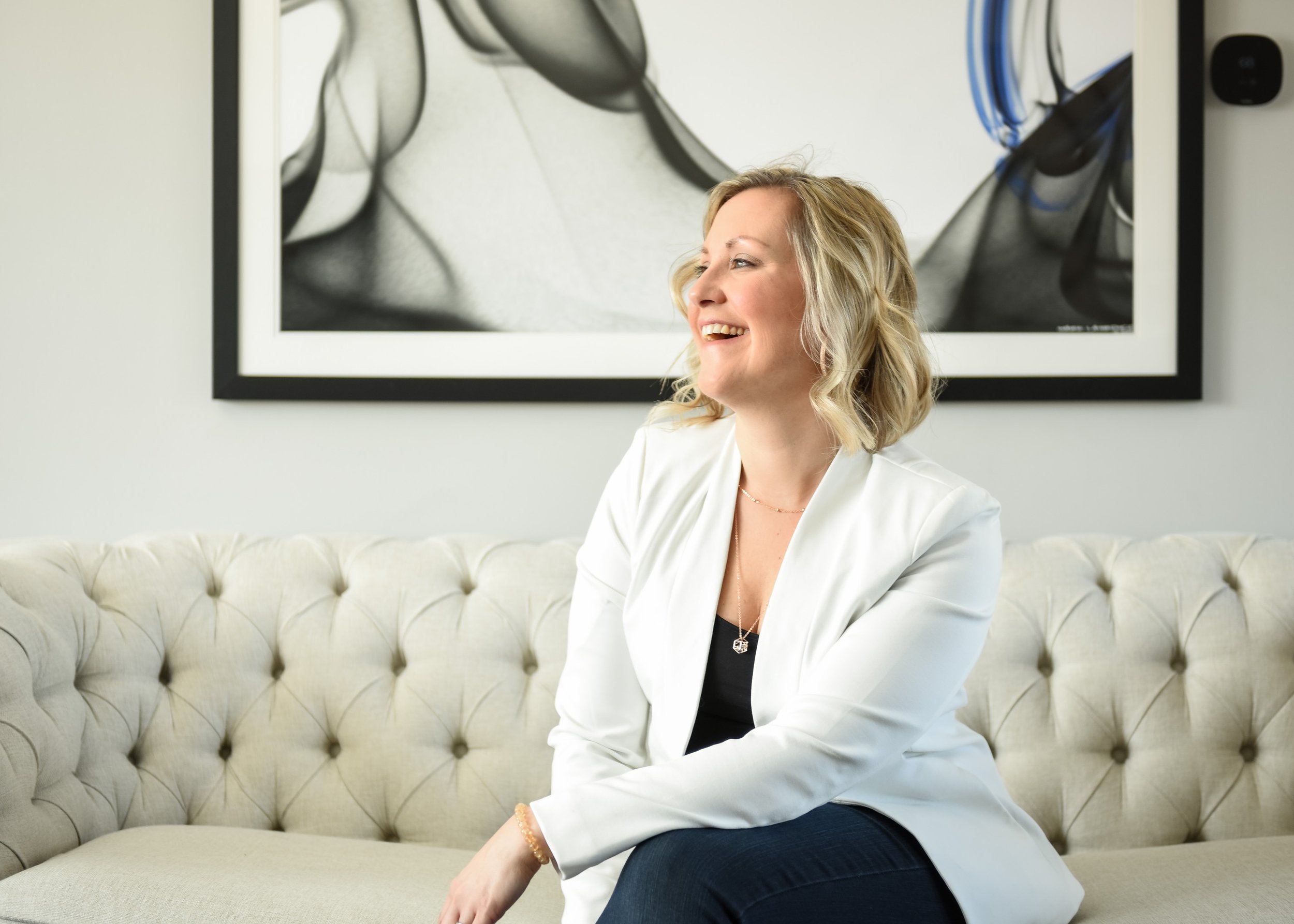 Woman in white blazer sitting on a tufted sofa, smiling and looking away - how to take personal branding photos for Instagram.