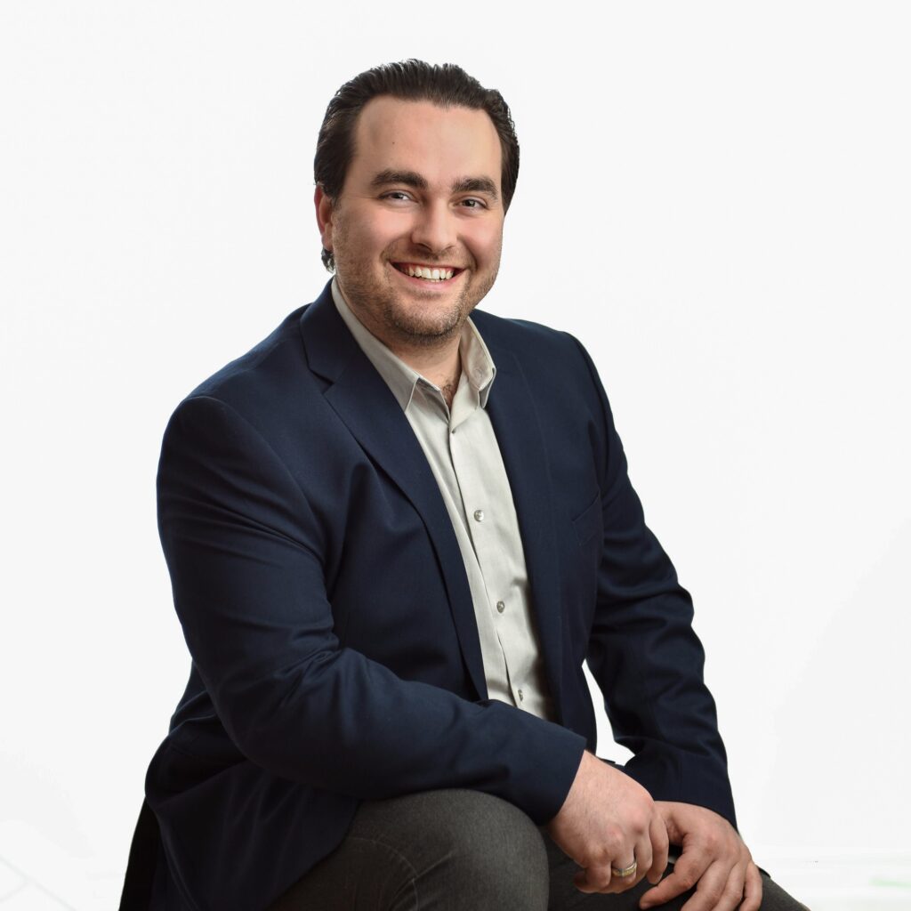  Professional man in a navy blazer and light shirt smiling confidently against a white background.