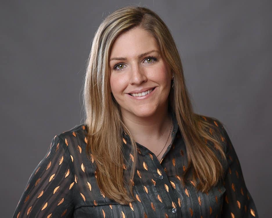 Professional corporate headshot of a woman with blonde hair wearing a black blouse with gold patterns, smiling warmly.