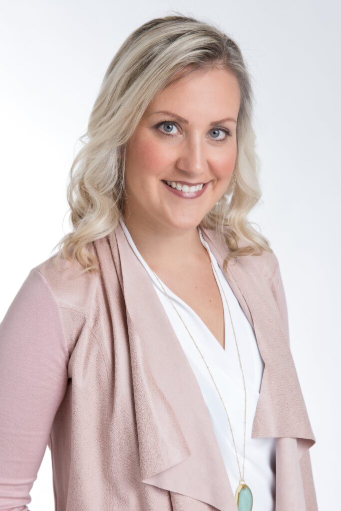 Professional corporate headshot of a blonde woman in a pink blazer, smiling brightly.