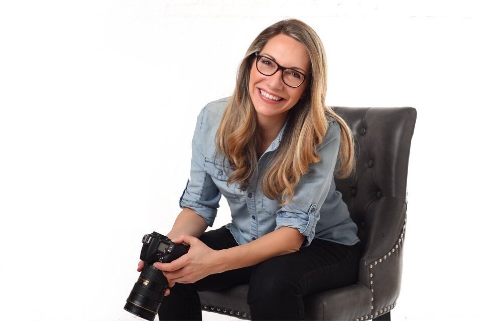 Carla Morgado photographer in Toronto holding a camera and smiling while sitting on a chair