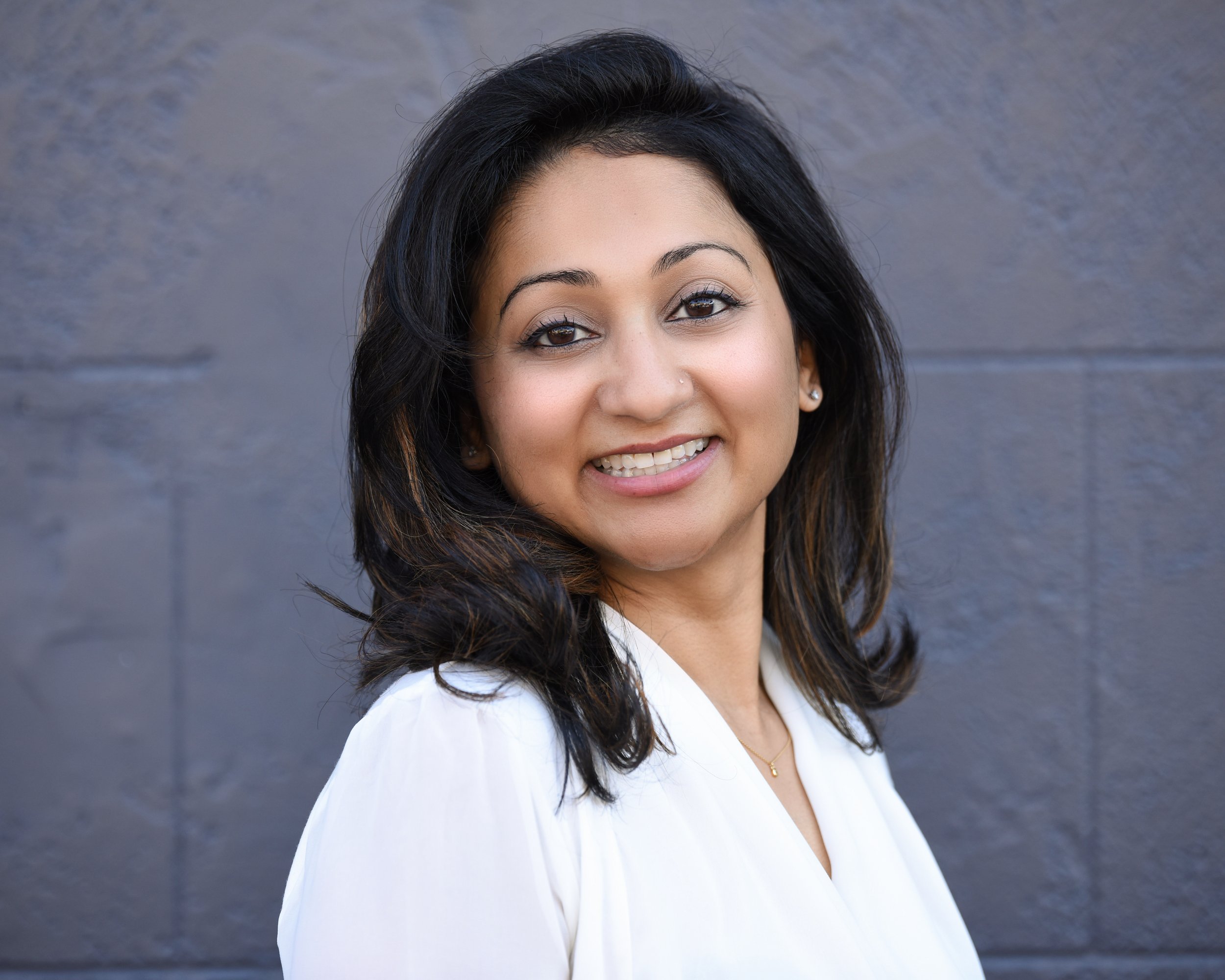 Smiling woman in a white blouse standing against a dark, textured background.