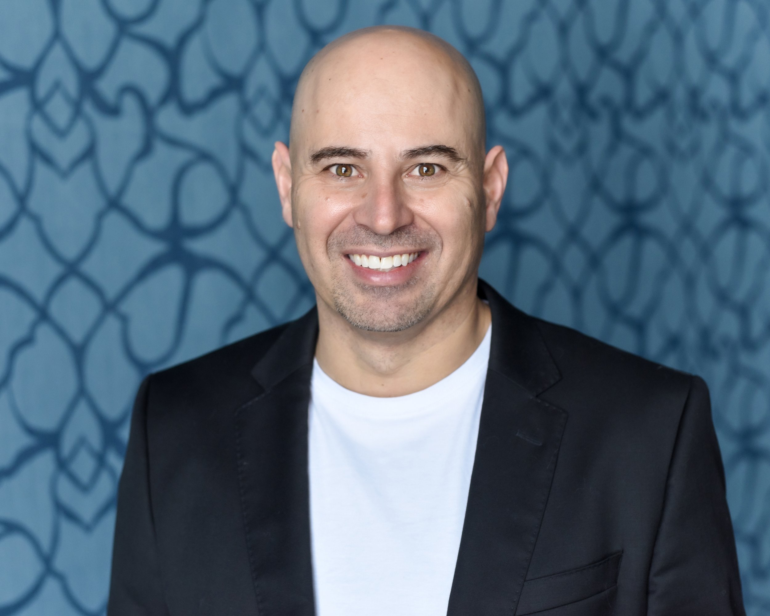 Smiling man in a black blazer with a white t-shirt against a patterned background.