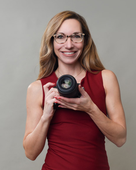 Smiling woman with glasses and long hair, holding a camera, wearing a red sleeveless top, posing against a gray background.