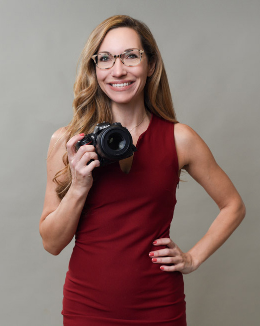 Woman with glasses and long hair, holding a camera and smiling, wearing a red sleeveless top, standing with one hand on her hip against a gray background.