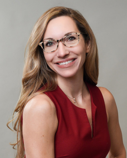 Professional headshot of a woman with glasses and long hair, wearing a red sleeveless top, smiling against a gray background.