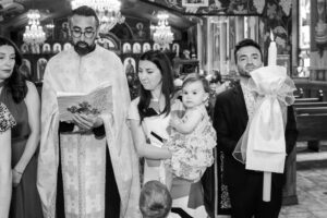 Priest reading from a book during a baptism ceremony while the family stands beside him, holding a baby.