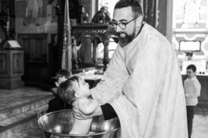 Priest performing a baptism by immersing a baby in a large metal basin inside a decorated church.