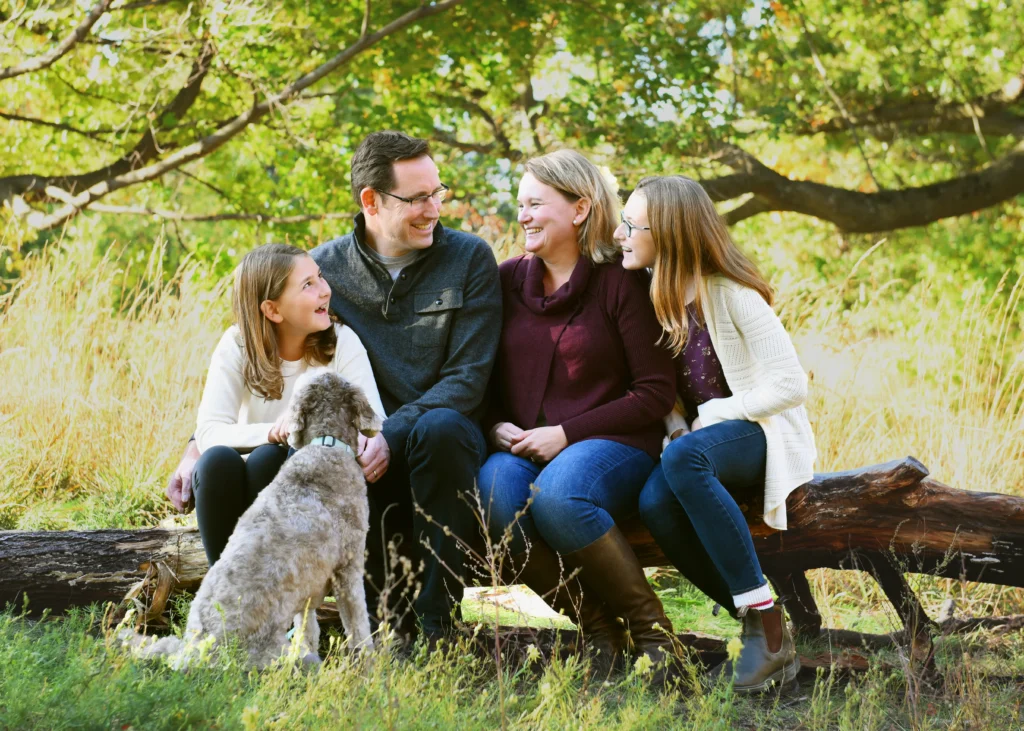 Family of four sitting on a log with their dog in a park, surrounded by autumn foliage.