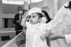 Baby in a white baptism outfit, held by a family member, adjusting their hat in a church setting.