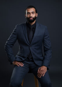 Young man sitting professionally in a dark background wearing a black suit and tie.