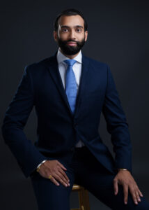 Young man sitting professionally in a dark background wearing a blue suit and tie.