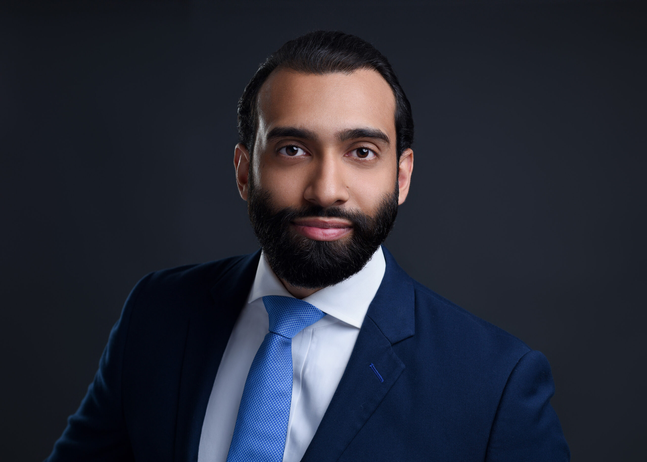 Close-up of a young man's professional headshot in a dark background wearing a blue suit and tie.