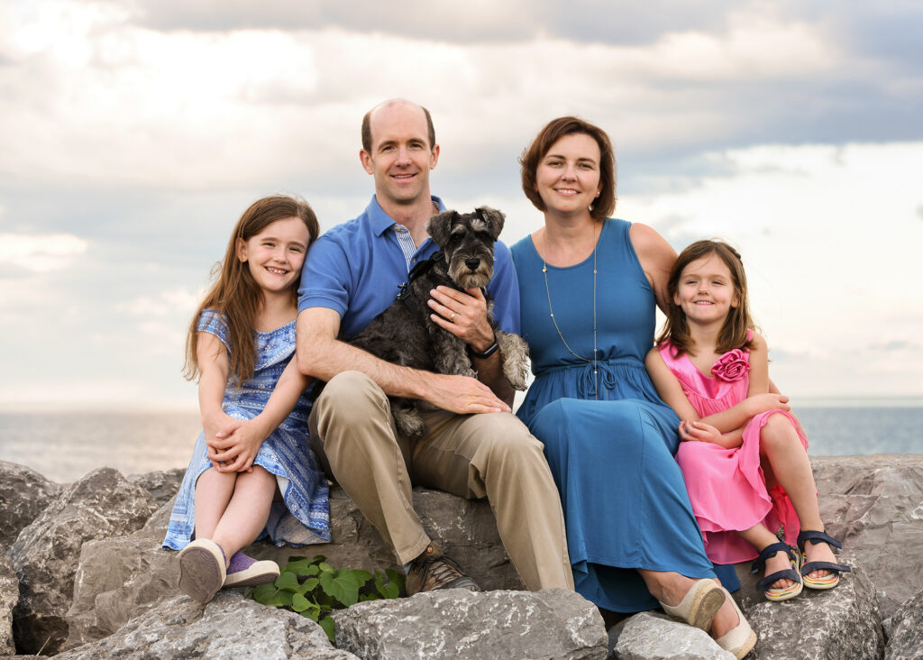 Family of four sitting on rocks by the lake with their dog, cloudy sky in the background.