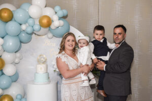 Family posing with their children in front of a decorative balloon arch and a baptism cake.