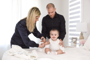 Godparents dressing their smiling baby in a baptism outfit in a bright and cozy room.