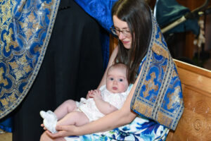 Mother holding her baby wrapped in a blue ceremonial cloth during a baptism in a church.