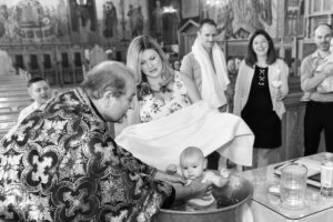 Priest performing a baptism by immersing a baby in a large metal basin inside a decorated church.