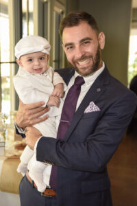 A man in a suit holds a baby boy in a white outfit, both smiling at the camera.