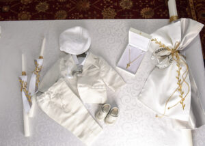 Baptism outfit and accessories including a white suit, hat, shoes, candles, and a cross necklace displayed on a table.