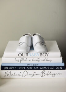 White baby shoes placed on a stack of personalized books announcing the birth of a baby boy.