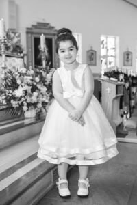 A young girl in a white dress stands next to a flower arrangement in a church.