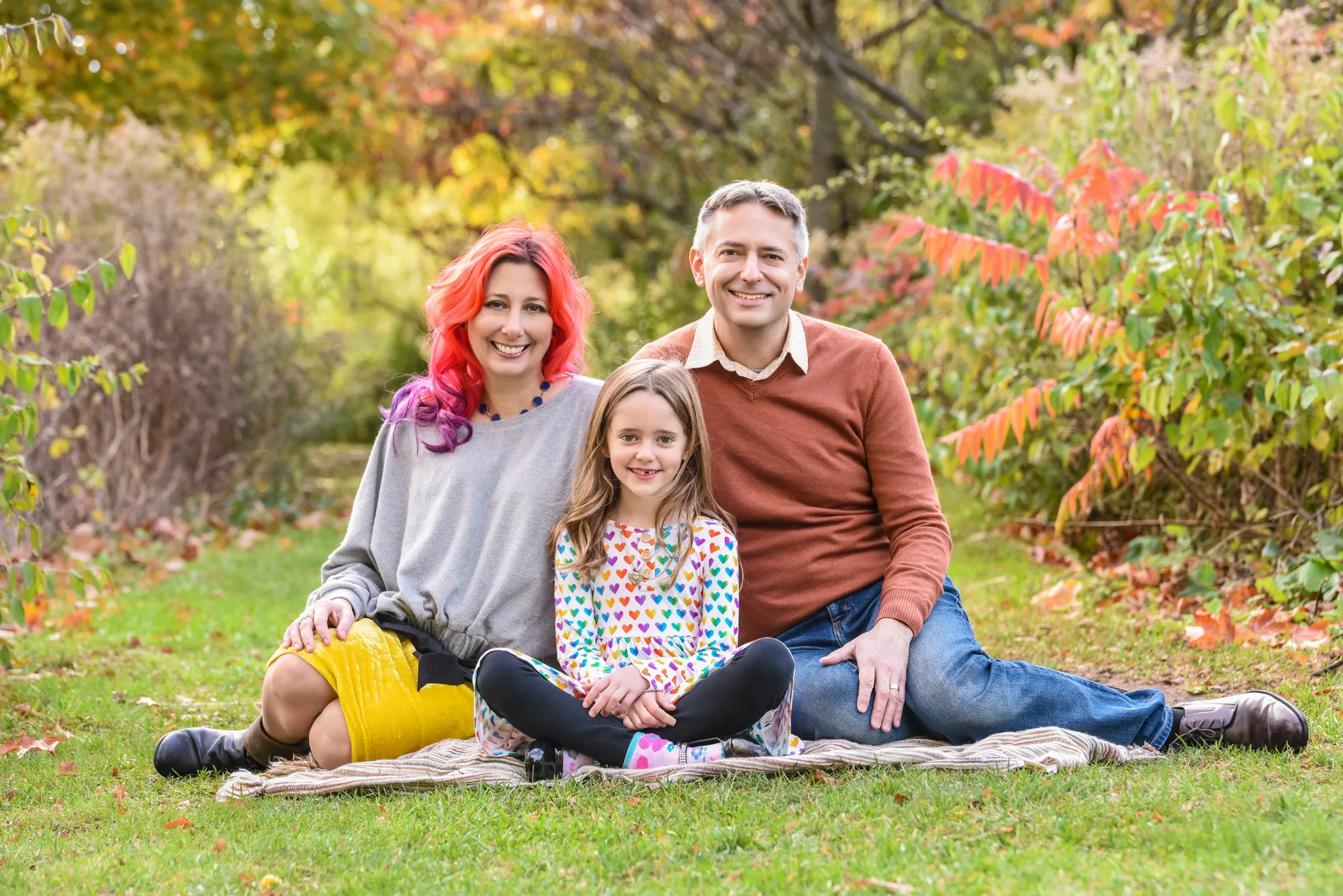 Family of three sitting on a blanket in a vibrant autumn park, smiling warmly at the camera.