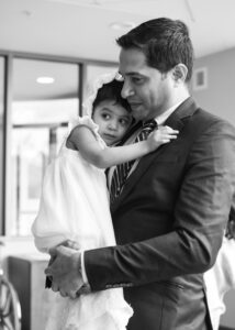 A man in a suit holds a young child dressed in white, both sharing a tender embrace.