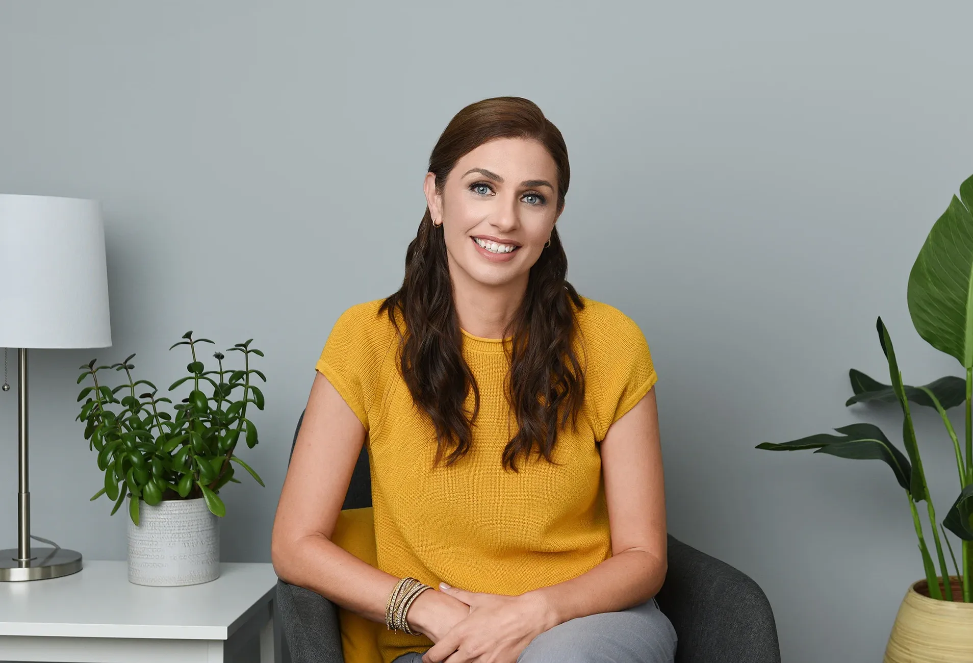 A professional headshot of a female psychotherapist sitting on a chair, smiling confidently, surrounded by plants, portraying a warm and approachable demeanor.