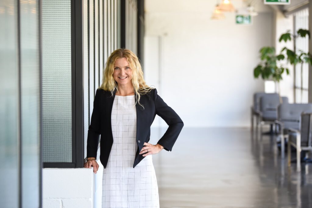 A businesswoman with blonde hair in a black blazer and white dress standing confidently in a modern office hallway.