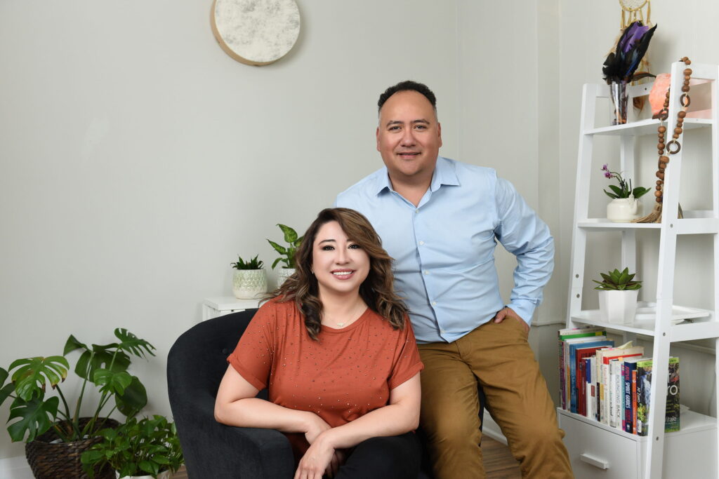 A professional man and woman sitting and standing in a home office environment, surrounded by plants and bookshelves.